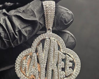 May include: A silver-toned pendant with the word "AYBEE" in a stylized font, set within a four-leaf clover shape. The pendant is encrusted with numerous small, sparkling stones, and has a matching bail for a chain.