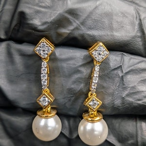 May include: A pair of gold-toned drop earrings. Each earring features a diamond-shaped top with small clear stones, a vertical line of clear stones, and a large, round, white pearl at the bottom. The earrings are displayed against a dark background.