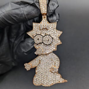 May include: A gold-colored pendant featuring a cartoon character design, encrusted with numerous small, sparkling stones. The pendant is suspended from a matching gold bail, and the character's face is detailed with black accents.