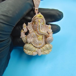 Puede incluir: Un colgante dorado con la forma de Ganesha, incrustado con numerosas piedras pequeñas y brillantes. El colgante presenta un diseño detallado de la deidad, con un tronco prominente y múltiples brazos. La fianza también está cubierta de piedras, creando un efecto ornamentado y lujoso.