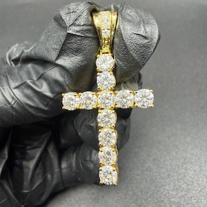 Pode incluir: Um pendente dourado em forma de cruz, incrustado com numerosas pedras preciosas redondas e transparentes. O pendente é apresentado sobre um fundo escuro, realçando os seus detalhes brilhantes. O design da cruz é um símbolo clássico de fé.