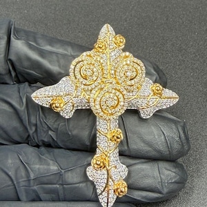 May include: A gold and silver cross pendant with a floral design. The cross features a detailed pattern of small, sparkling stones and gold accents. The cross is held against a dark background.
