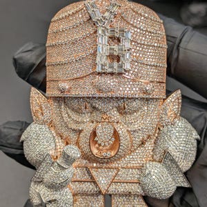May include: A large, ornate pendant featuring a figure design, encrusted with numerous small, sparkling diamonds. The pendant has a rose gold tone with a silver-colored letter "B" at the top. The figure has detailed features and is held by a gloved hand.