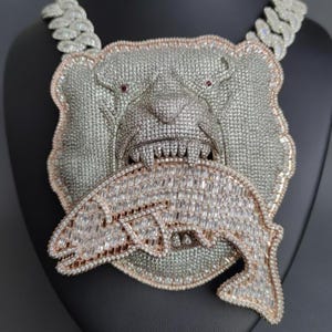 May include: A silver-toned pendant necklace featuring a bear head design with red eyes, biting a fish. The bear and fish are encrusted with sparkling stones. The necklace has a thick, silver chain.
