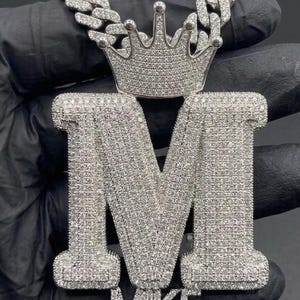 May include: A silver pendant necklace with a large letter "M" and the name "MIKE" below. The pendant is covered in small, sparkling stones and has a crown on top. The chain is also silver.