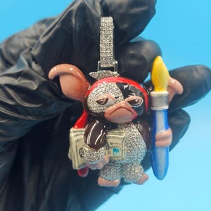 May include: A detailed pendant featuring a cartoon character with a red and black hat, holding a paintbrush and money. The pendant is encrusted with small, sparkling stones and has a silver-coloured bail.