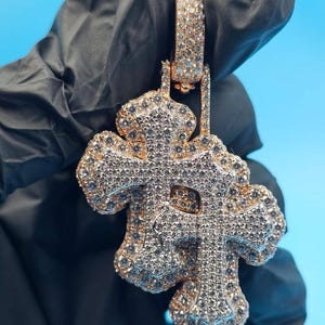 May include: A gold-toned pendant in the shape of a cross, encrusted with numerous small, sparkling gemstones. The pendant has a detailed design with multiple layers and a bail for attaching to a chain. The gemstones are set in a pavé style.