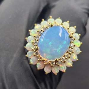 May include: A gold ring featuring a large, oval, light blue opal gemstone at the center, surrounded by smaller opals and diamonds. The ring has a floral design, with the smaller opals forming a halo around the central stone.