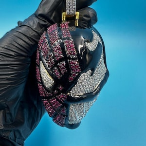 May include: A pendant with a black and silver design and red accents. The pendant has a silver bail and is encrusted with sparkling stones. The design is a stylized mask with a black glossy finish and a silver and red stone pattern.