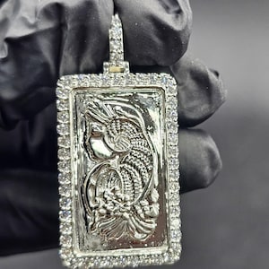 Puede incluir: Colgante rectangular plateado con un diseño en relieve detallado y un borde de pequeñas piedras transparentes. El colgante tiene una argolla en la parte superior para una cadena. El diseño interior es un relieve detallado de una figura.