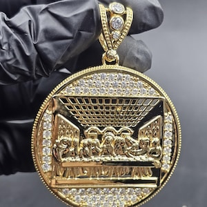 May include: A gold pendant depicting the Last Supper, with intricate detailing. The round pendant is embellished with small, clear gemstones around the edge and on the bail. The image is held by a gloved hand.