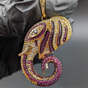 Könnte beinhalten: Goldfarbener Anhänger in Form eines Elefantenkopfes, besetzt mit roten und weißen Edelsteinen. Der Anhänger hat ein detailliertes Design mit einem eingerollten Rüssel und einer Schlaufe für eine Kette. Das Auge des Elefanten ist ein einzelner, klarer Edelstein.