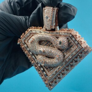 May include: A diamond-encrusted pendant in a rose gold setting. The pendant features a snake design within a diamond-shaped frame, with intricate detailing and sparkling stones. The background is a solid blue.