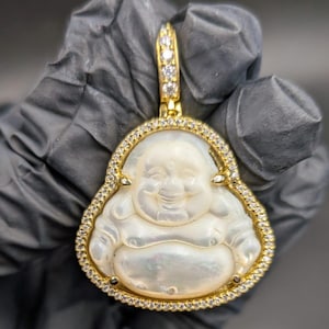 May include: A gold-colored pendant featuring a smiling Buddha figure carved from a pearlescent material. The pendant is outlined with small, clear gemstones and has a bail adorned with additional gemstones.