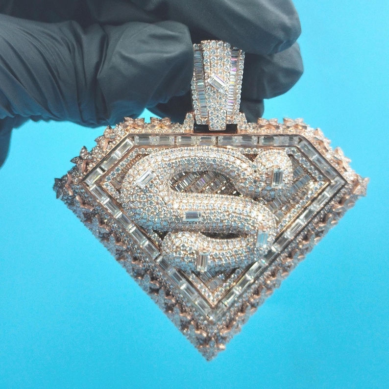 May include: A diamond-encrusted pendant in the shape of a diamond, featuring the letter "S" in the center. The pendant is rose gold in color and is covered in small, sparkling diamonds and larger baguette-cut diamonds.