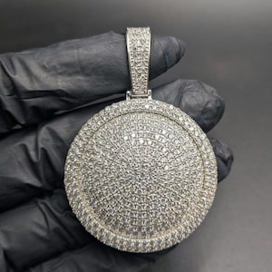May include: A round, silver-colored pendant encrusted with numerous small, clear gemstones. The pendant has a bail at the top for a chain. The gemstones cover the entire surface of the pendant, creating a sparkling effect. The pendant is held by a gloved hand.