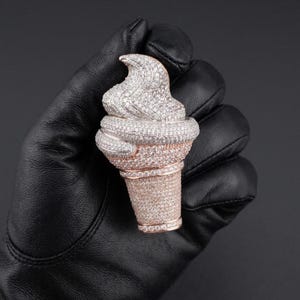 May include: A diamond-encrusted ice cream cone pendant. The cone is rose gold, and the ice cream is covered in white diamonds. The pendant is held in a black leather glove against a dark background.