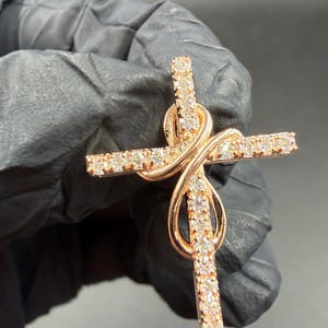 May include: Rose gold cross pendant with an infinity symbol design. The cross is encrusted with small, sparkling clear stones. The pendant is held by a gloved hand against a dark background.