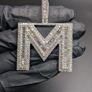 May include: A diamond-encrusted pendant in the shape of the letter "M". The pendant is made of silver-coloured metal and is covered in small, sparkling diamonds. The pendant is held against a black background.
