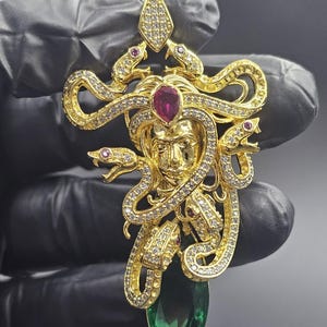 Puede incluir: Un colgante dorado con un diseño de Medusa. El colgante presenta una cara central con serpientes, adornada con piedras preciosas claras y rojas. Una gran piedra verde en forma de lágrima cuelga debajo, contrastando con el oro y las piedras preciosas.