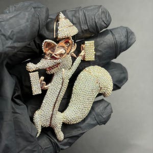 Puede incluir: Un colgante detallado con un diseño de ardilla de dibujos animados. El colgante está incrustado con pequeñas piedras brillantes, con una combinación de colores oro rosa y plata. La ardilla sostiene dos objetos rectangulares y tiene un sombrero en forma de árbol de Navidad.