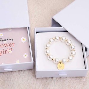 May include: Two white gift boxes, one open to reveal a pink card with the text "will you be my flower girl?" and daisy illustrations. The other box contains a pearl bracelet with a gold charm.