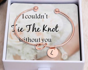 May include: Rose gold-colored bracelet with a knot design and a small circular charm with the letter "L". The bracelet is presented in a white gift box with the text "I couldn't Tie The Knot without you".