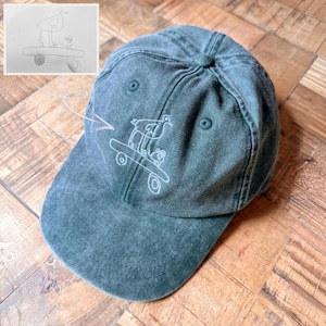 May include: A dark green baseball cap featuring a white embroidered line drawing of a bird standing on a skateboard next to a small mouse. The design is inspired by the sketch shown in the upper left corner of the image, which depicts the same scene.