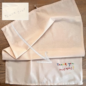 May include: A folded off-white cotton tea towel featuring a white embroidered arrow pointing towards colourful text that reads Thank you mrs ayres! A separate inset shows the handwritten text Thank you! in pencil on a white background.