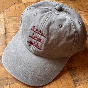 May include: Grey baseball cap with a curved peak and a button on top. The cap has the embroidered text "Gaga Loves Golf" in red thread. The hat is made of a soft, textured fabric.