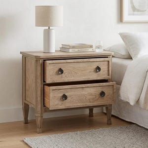 Op de afbeelding: Een nachtkastje van licht eikenhout met twee lades, waarvan één open. Het nachtkastje heeft zwarte, ronde ladeknopen en staat naast een bed met wit beddengoed. Een witte lamp met een beige kap staat erop.
