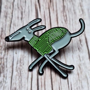 Puede incluir: Un pin de esmalte gris de un galgo corriendo con un suéter verde. El perro tiene un hocico largo y orejas grandes. El pin tiene contornos negros y está sobre un fondo de vetas de madera.