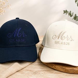 May include: A navy blue baseball cap with the embroidered text Mr. EST. 4.9.26 and a white baseball cap with the embroidered text Mrs. EST. 4.9.26. Both caps feature a structured crown, a curved brim, and matching tonal stitching throughout the design.