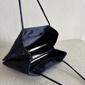 Peut inclure: Un sac &agrave; main en satin bleu marine fonc&eacute; avec une finition lisse et brillante et de fines bretelles. Le sac est ouvert, r&eacute;v&eacute;lant un int&eacute;rieur en tissu assorti avec un compartiment central zipp&eacute; et une poche, contenant un objet blanc.