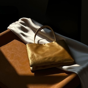 Peut inclure: Un sac &agrave; main rectangulaire en satin dor&eacute; dot&eacute; d'une anse unique sur le dessus. Le sac pr&eacute;sente une finition lisse et lustr&eacute;e qui refl&egrave;te la lumi&egrave;re, cr&eacute;ant des reflets et des ombres sur sa surface. Le design est simple, structur&eacute; et compact.