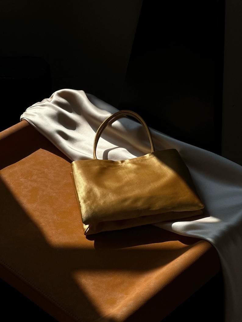 Peut inclure: Un sac &agrave; main rectangulaire en satin dor&eacute; dot&eacute; d'une anse unique sur le dessus. Le sac pr&eacute;sente une finition lisse et lustr&eacute;e qui refl&egrave;te la lumi&egrave;re, cr&eacute;ant des reflets et des ombres sur sa surface. Le design est simple, structur&eacute; et compact.