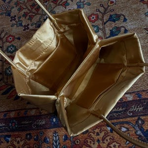 Peut inclure: Un sac &agrave; main en satin dor&eacute; avec une ouverture rectangulaire structur&eacute;e et deux fines bandouli&egrave;res assorties. L'int&eacute;rieur est doubl&eacute; d'un tissu dor&eacute; lisse et comporte deux poches rectangulaires ouvertes sur les parois lat&eacute;rales internes.