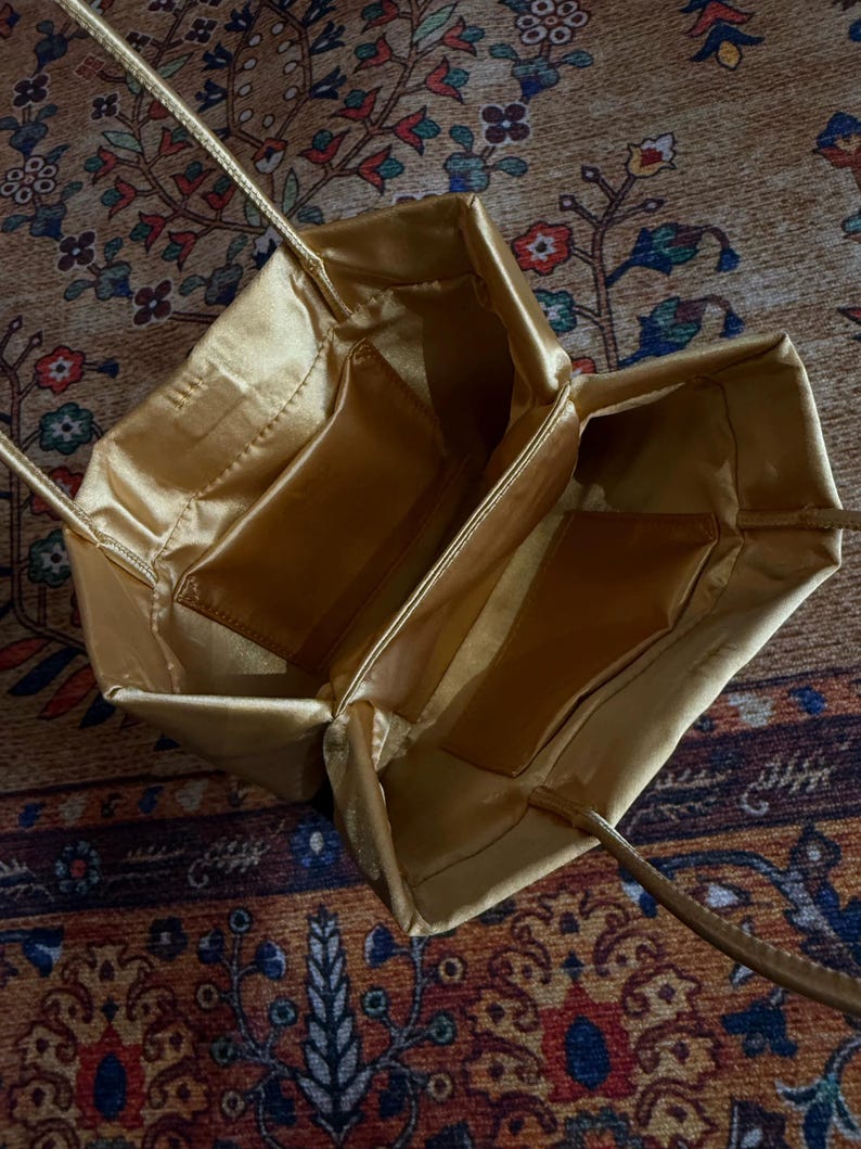 Peut inclure: Un sac &agrave; main en satin dor&eacute; avec une ouverture rectangulaire structur&eacute;e et deux fines bandouli&egrave;res assorties. L'int&eacute;rieur est doubl&eacute; d'un tissu dor&eacute; lisse et comporte deux poches rectangulaires ouvertes sur les parois lat&eacute;rales internes.