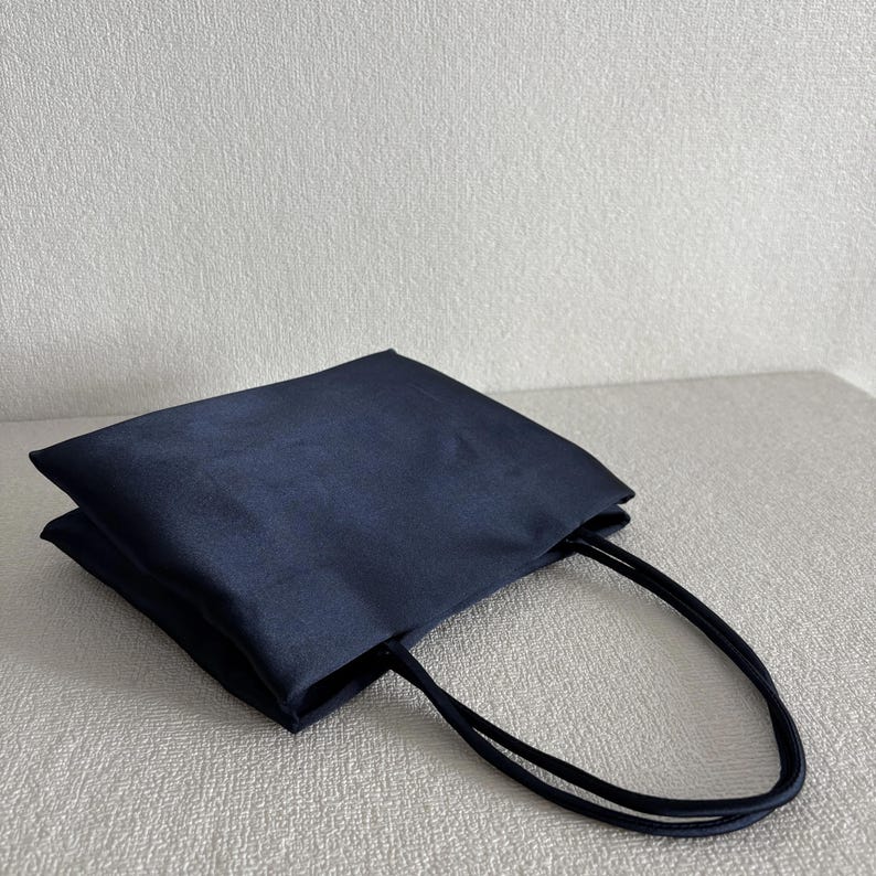 Peut inclure: Un sac fourre-tout bleu marine fonc&eacute; avec une finition lisse et satin&eacute;e. Le sac pr&eacute;sente une structure rectangulaire &agrave; fond plat et deux fines anses assorties bleu marine fonc&eacute; fix&eacute;es au bord sup&eacute;rieur du compartiment principal.