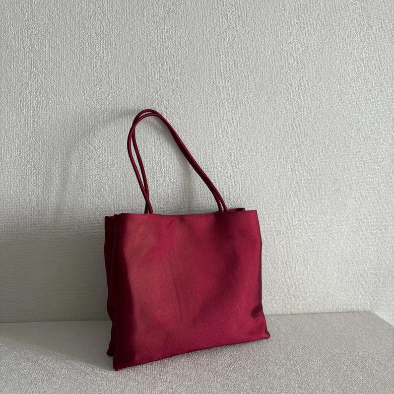 Peut inclure: Un sac fourre-tout rectangulaire rouge fonc&eacute; fabriqu&eacute; dans un tissu lisse semblable &agrave; du satin. Le sac pr&eacute;sente deux fines bretelles assorties fix&eacute;es au bord sup&eacute;rieur, offrant une silhouette simple et structur&eacute;e avec un l&eacute;ger &eacute;clat.