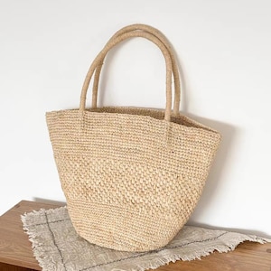 May include: A woven straw tote bag with two rounded handles. The bag is a natural beige color and has a textured appearance. The bag is sitting on a wooden surface with a fringed linen cloth.