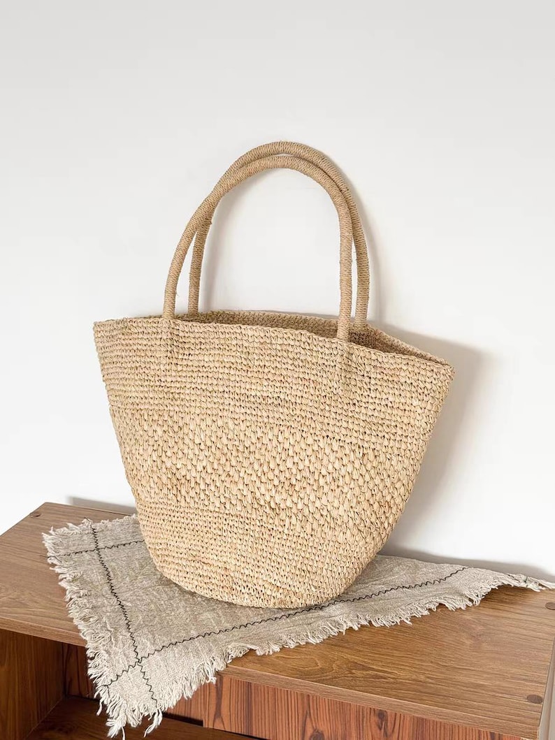 May include: A woven straw tote bag with two rounded handles. The bag is a natural beige color and has a textured appearance. The bag is sitting on a wooden surface with a fringed linen cloth.
