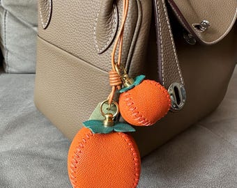 Peut inclure: Un sac à main en cuir avec un charm orange décoratif. Le charm est en cuir orange avec des accents de feuilles vertes et un fermoir doré. Le sac à main est de couleur neutre avec des coutures blanches.