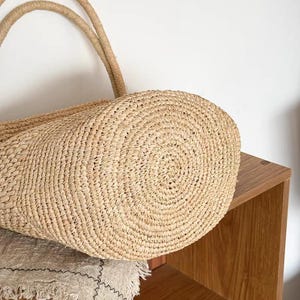 May include: A close-up of a natural woven straw tote bag with two rope handles. The bag is light tan in color and has a circular base. The bag is sitting on a beige linen cloth.