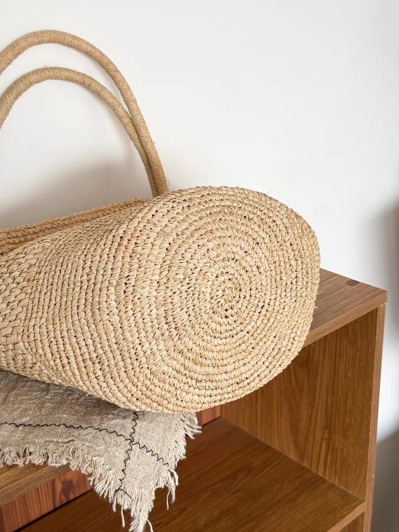 May include: A close-up of a natural woven straw tote bag with two rope handles. The bag is light tan in color and has a circular base. The bag is sitting on a beige linen cloth.