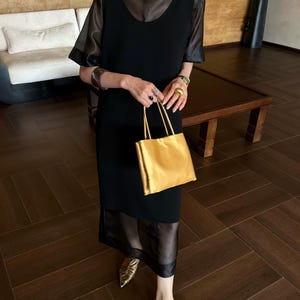 Peut inclure: Une robe midi noire sans manches avec une superposition transparente &agrave; manches courtes et un col haut. La tenue est assortie d'un sac &agrave; main rectangulaire dor&eacute; m&eacute;tallis&eacute;, de chaussures plates dor&eacute;es &agrave; bout pointu, d'un large bracelet manchette sombre et de bagues dor&eacute;es.