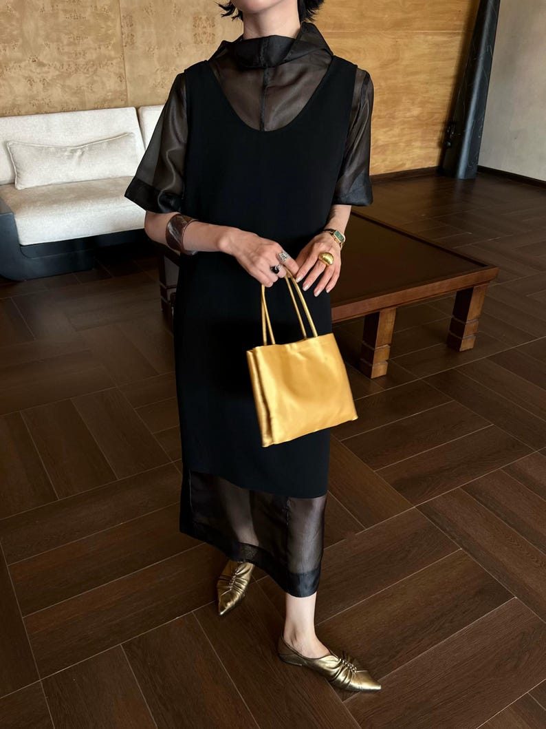 Peut inclure: Une robe midi noire sans manches avec une superposition transparente &agrave; manches courtes et un col haut. La tenue est assortie d'un sac &agrave; main rectangulaire dor&eacute; m&eacute;tallis&eacute;, de chaussures plates dor&eacute;es &agrave; bout pointu, d'un large bracelet manchette sombre et de bagues dor&eacute;es.