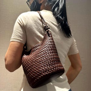 Peut inclure: Un sac à bandoulière en cuir marron tressé avec une bandoulière tressée. Le sac présente un motif texturé unique et un détail noué en haut de la bandoulière. Le sac est porté à l'épaule.