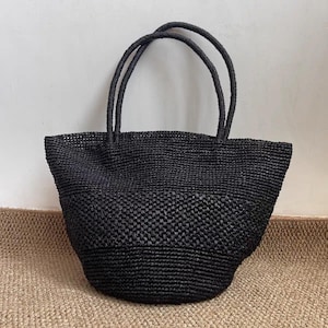 May include: A black woven tote bag with two rounded handles. The bag has a structured shape and a textured surface. The tote bag is made of a dark material and is suitable for carrying various items.