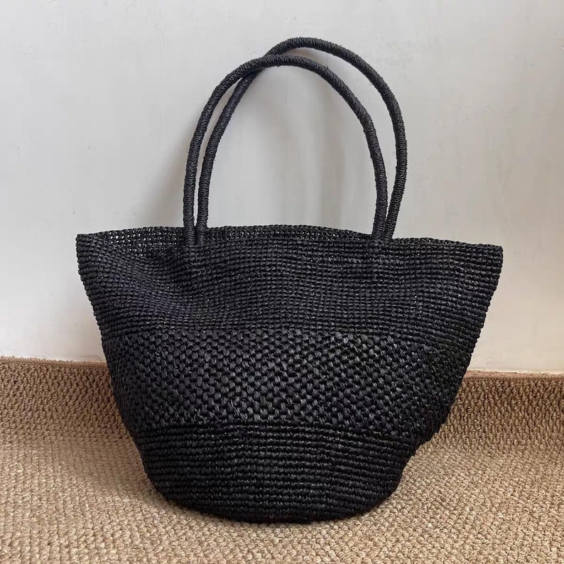 May include: A black woven tote bag with two rounded handles. The bag has a structured shape and a textured surface. The tote bag is made of a dark material and is suitable for carrying various items.